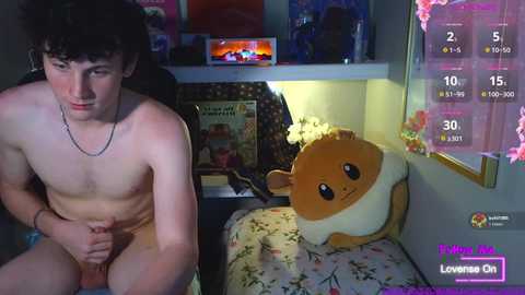 A young, nude man with curly hair sits on a bed in a dimly lit room. A plush Eevee toy is beside him. The background includes posters and a TV.