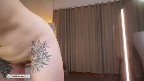 A close-up video of a light-skinned person's side, revealing a detailed black and grey floral tattoo on the left ribcage. The background features beige curtains and a warm, soft light.