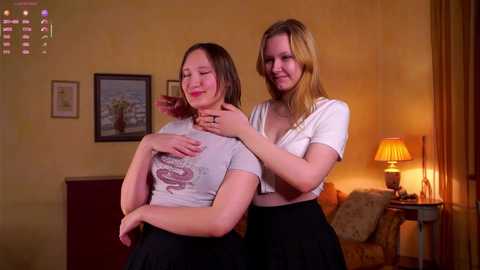 Video of two young women, one with shoulder-length hair and another with long blonde hair, in a warmly lit living room, both smiling.