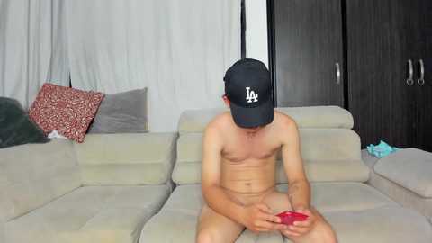 Video of a young, slender, nude Asian man with a small penis, wearing a black cap, seated on a beige sofa in a minimalist living room, holding a red smartphone.