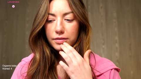 Video of a young woman with long, wavy brown hair, wearing a pink jacket, touching her hair thoughtfully against a textured, muted gray background.