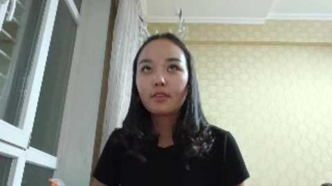 Video of an Asian woman with long black hair and wearing a black shirt, standing in a room with yellow wallpaper and a window to the left.