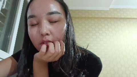 A video of an Asian woman with long black hair and light skin, wearing a black top, licking her fingers with eyes closed, in a beige-patterned room.