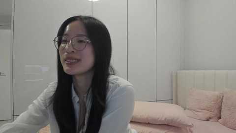 Video of a smiling East Asian woman with long black hair, wearing glasses, a light blue shirt, and a pink blanket, sitting on a bed in a minimalist, white-walled bedroom.