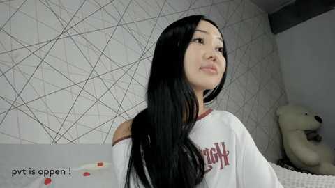 Video of an Asian woman with long black hair, fair skin, and a slender physique, wearing a white off-shoulder t-shirt with red text, standing against a geometric-patterned wall, holding a teddy bear.