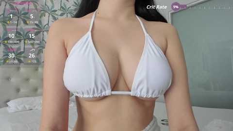 Video of a woman with fair skin, wearing a white bikini top, revealing ample cleavage and a toned midsection. Background features a bed with white sheets and a floral-patterned wall.