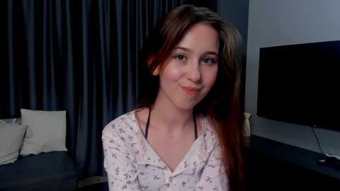 Video of a young woman with long, light brown hair, wearing a white pajama shirt, smiling, in a dimly lit room with dark curtains and a flat-screen TV.