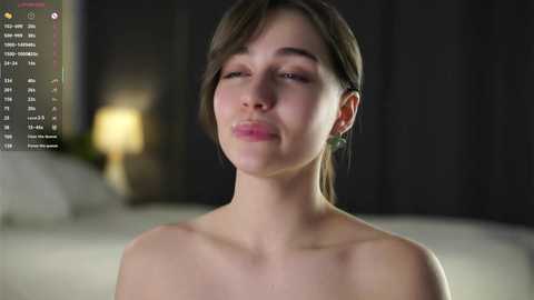 A video of a topless young woman with light skin, brown hair, and green earrings, in a dimly-lit bedroom with blurred background.