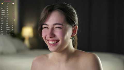A video of a smiling East Asian woman with straight black hair, wearing a strapless top, in a dimly-lit room with blurred furniture.