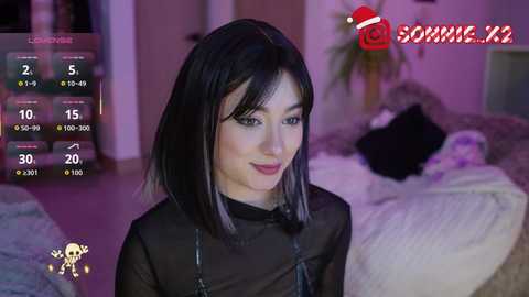 A video of a young woman with straight black hair and a black sheer top, set in a dimly-lit bedroom. The background shows a bed with white and black bedding.