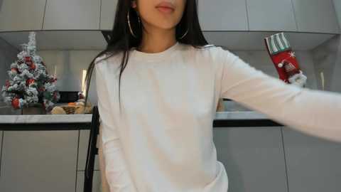 Video of a woman with long black hair, wearing a white long-sleeve shirt, holding a red and silver can. Background features a modern kitchen with white cabinets and a snow-dusted Christmas tree.