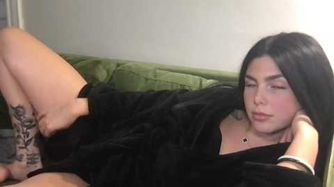 Video of a light-skinned woman with long black hair, wearing a black velvet dress, lying on a green couch, with a tattoo on her thigh.