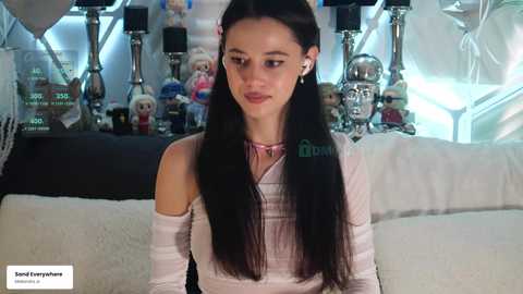 Video of a slender young woman with long black hair, wearing a white off-shoulder top, sitting in a modern room with white furniture, surrounded by toy figurines.