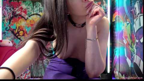 Video of a young woman with long, straight black hair, wearing a strapless purple top and black choker, leaning on her elbow, against a vibrant, colorful, abstract wall with graffiti art.