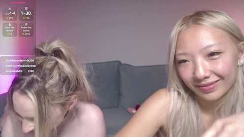 A video of two nude young women with fair skin, one blonde with a ponytail, the other with long hair, smiling and lying on a couch in a dimly lit room.