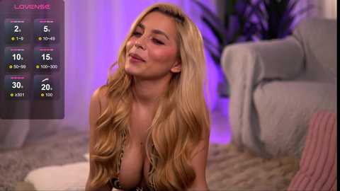 A video of a blonde woman with long, wavy hair and large breasts in a low-cut top, smiling seductively in a cozy living room with a grey couch and purple lighting.