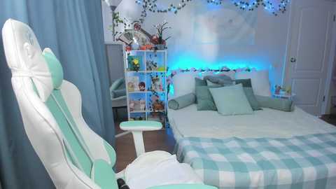 A video of a modern, brightly lit bedroom with a white gaming chair, a white desk, and a white bed adorned with pastel green and blue bedding. Fairy lights and plants decorate the walls.