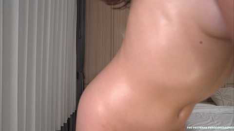 Video of a nude woman's side profile, highlighting smooth, light-toned skin and small breasts, in a bedroom with vertical blinds and a bed visible in the background.