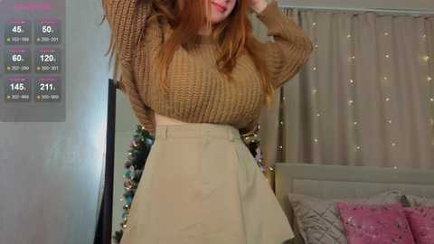 Video of a fair-skinned, red-haired woman in a brown knitted sweater and high-waisted beige skirt, standing in a cozy bedroom with a Christmas tree, bed, and window with sheer curtains.