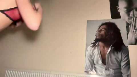 Video of a young man with long, dark hair, wearing a light blue shirt, standing against a beige wall with a poster of a black man in the background.