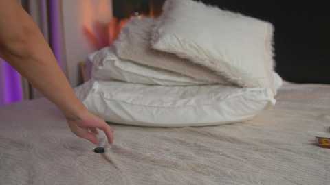 Video of a person's hand smoothing white pillow stuffing onto a fluffy white pillow, with a background of white sheets and a dark wall.