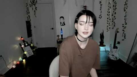 Video of a pale woman with dark hair, wearing a brown shirt and black choker, in a dimly lit room with fairy lights, hanging plants, and a white door in the background.