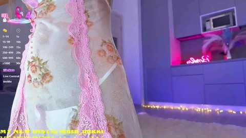 Video of a woman in a pastel floral dress with pink lace trim, standing in a dimly lit room with purple and pink lighting.