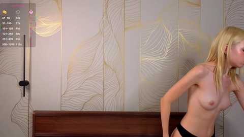 Media: Video of a nude, slender, fair-skinned woman with blonde hair, wearing only black panties, standing in a room with a light beige wall and leaf-patterned wallpaper.