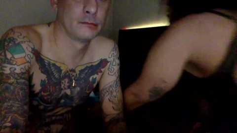 Video of a pale-skinned, tattooed man with short hair, wearing a colorful, sleeveless shirt, sitting on a bed. Another person, partially visible, is in the background.