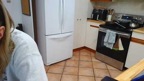 Video of a woman in a white robe in a small kitchen with a white refrigerator, black oven, tiled floor, and wooden cabinets.