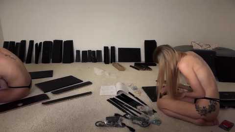 Media: Video of a blonde, tattooed woman kneeling on a beige carpet, assembling black speaker components, surrounded by various electronic parts and tools, in a cluttered room with off-white walls.