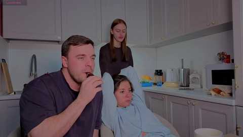 A video depicts a man in a dark shirt eating, a woman in a black top, and a child in a blue gown in a modern kitchen with white cabinets and appliances.