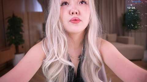 A video of an Asian woman with long, platinum blonde hair, wearing a black sleeveless top, looking pensive in a modern, softly lit living room with beige curtains and a white sofa.