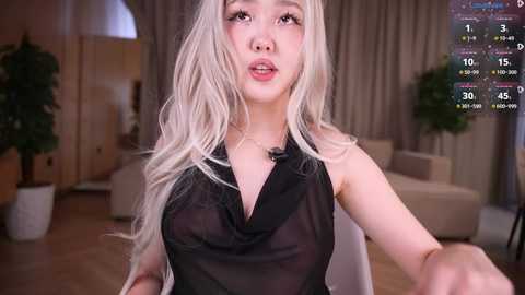Video of a young Asian woman with long, platinum blonde hair, wearing a sheer black dress, sitting in a modern living room with beige curtains and potted plants.