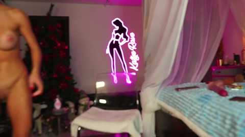 Video of a woman, topless with medium breasts, standing in a bedroom with a neon silhouette of a woman on the wall. Pink lighting, bed with blue blanket, white chair, and a Christmas tree in the background.