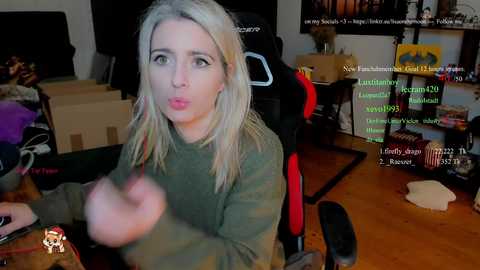 Video of a blonde woman in a green sweater, making a surprised face, surrounded by gaming equipment and cardboard boxes in a cluttered room.