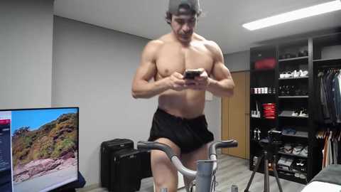 Video of a muscular shirtless man wearing black shorts, standing in a gym with a flat-screen TV displaying a beach scene, cycling equipment, and a bookshelf in the background.