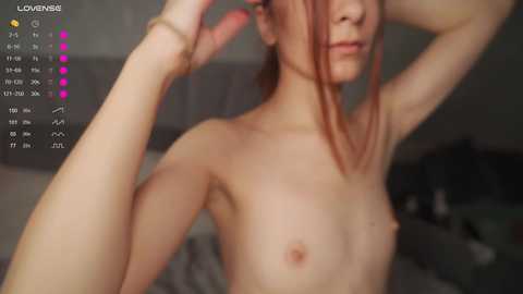 Video of a slender, pale-skinned, topless woman with small breasts and red hair, adjusting her hair, in a dimly lit room with a calendar displaying \"LOVENES\" in the corner.