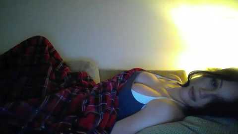 A video shows a young woman with long brown hair, wearing a red plaid shirt and a white bra, lying on a couch under a plaid blanket, illuminated by a yellow lamp.