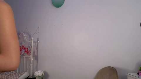A video of a bare-chested man in a pastel-colored room with a white metal bed frame, a green balloon, and a white chair.