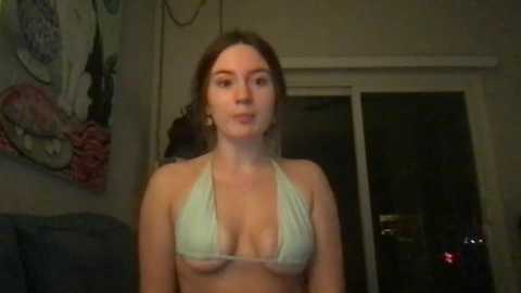 A video of a young woman with light skin and medium-length brown hair, wearing a revealing light green bikini top, standing in a dimly lit room with a floral painting on the wall.