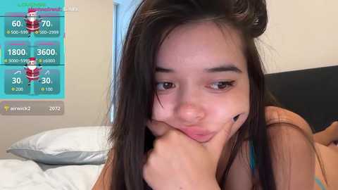 A video of a young Asian woman with long black hair, resting her chin on her hand, looking slightly melancholic. She is wearing a brown top. The background shows a bed with white sheets and a black headboard.