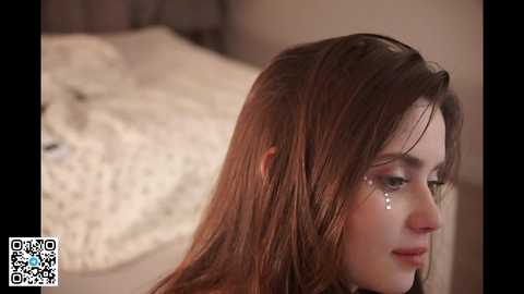 Video of a young woman with long, wet auburn hair, tear-streaked makeup, and a pensive expression. She sits on a beige bedspread in a softly lit room.
