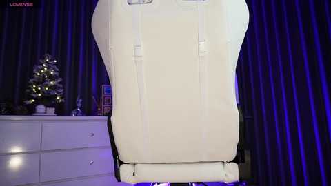 Video of a cream-colored gaming chair with adjustable armrests, set against dark purple curtains. A small decorated Christmas tree with white lights and ornaments is on a white dresser to the left.