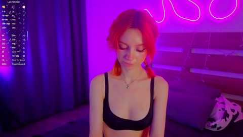 Video of a young woman with red hair, wearing a black bra, standing in a dimly lit room with purple neon lights, a cow print pillow, and a digital camera display on the left.