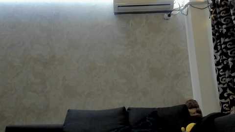 A video of a beige marble wall with a black leather couch featuring a black and yellow pillow. An air conditioner is mounted above, and a black and white patterned curtain is partially visible on the right.