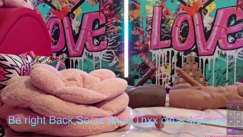 Video of a person lying on a pink, fluffy bedspread, surrounded by vibrant, graffiti-covered walls with the word \"LOVE\" in large, bold letters. Text overlays: \"Be right back, some snail oil at my neck.\