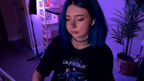 Video of a young woman with blue hair, wearing a black Slayer T-shirt, choker, and white earbuds, standing indoors with purple lighting, plants, and a shelf in the background.