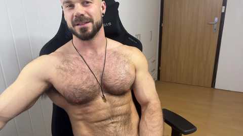 Video of a muscular, bearded, fair-skinned man with a hairy chest, sitting shirtless in a black gaming chair, wearing a pendant necklace, in a wooden-floored room with white paneling and a door.