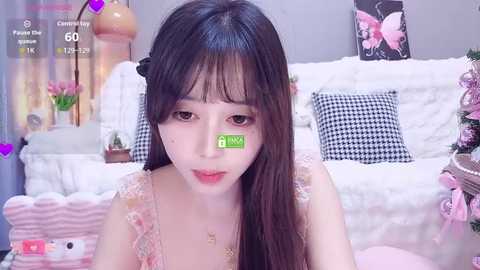 A video of an Asian woman with long black hair and fair skin, wearing a pink lace dress, sitting on a white bed with pink pillows and floral decor.